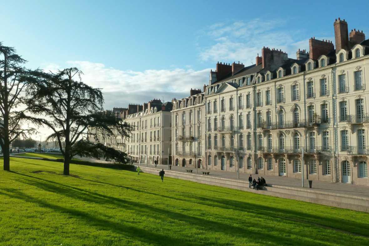 Façade d'immeubles de type nantais en pierre de taille longeant un quai avec une pelouse et un grand arbre au premier plan