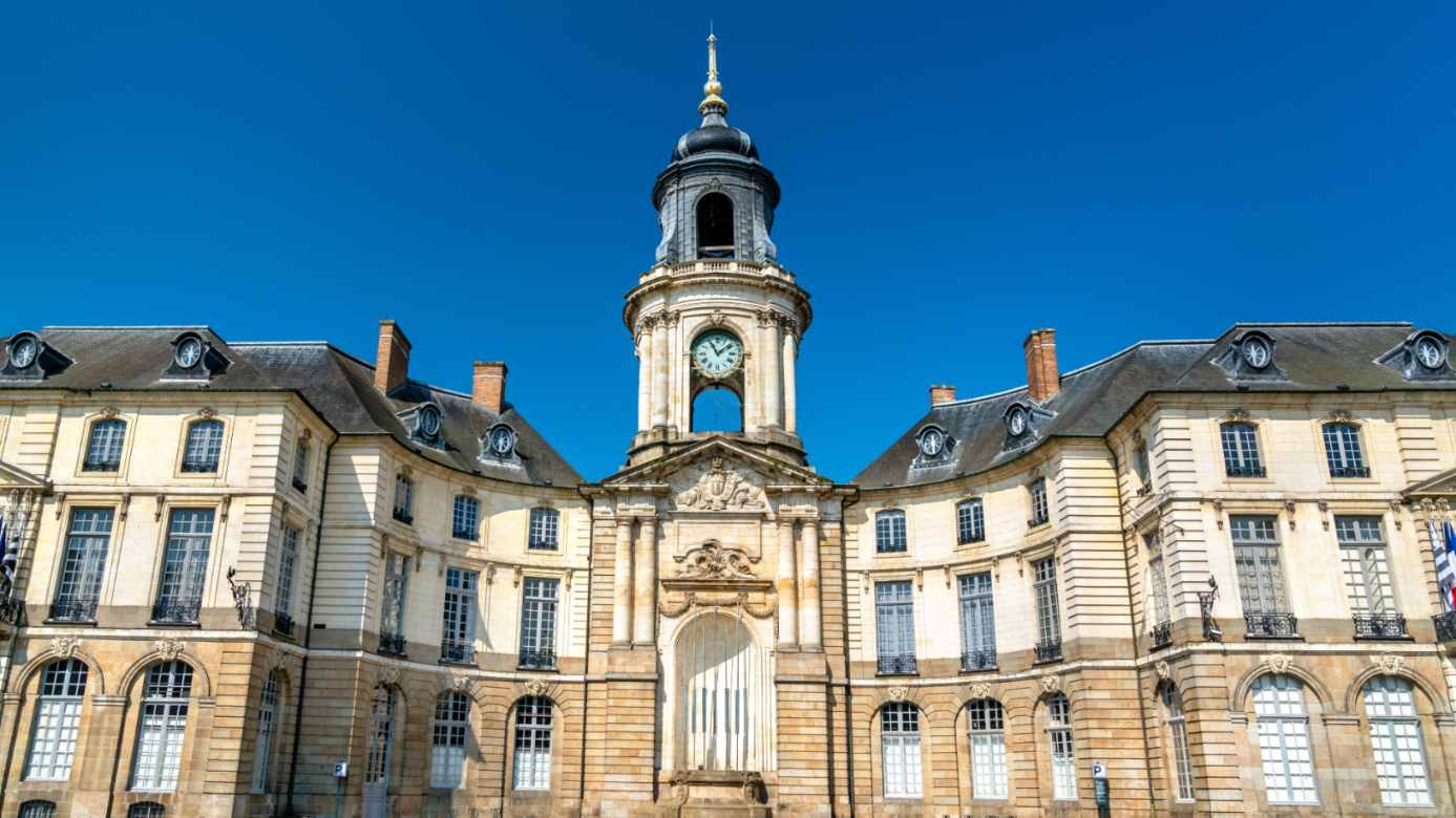 Façade symétrique en pierre de taille avec horloge centrale et toit en ardoise sous un ciel bleu