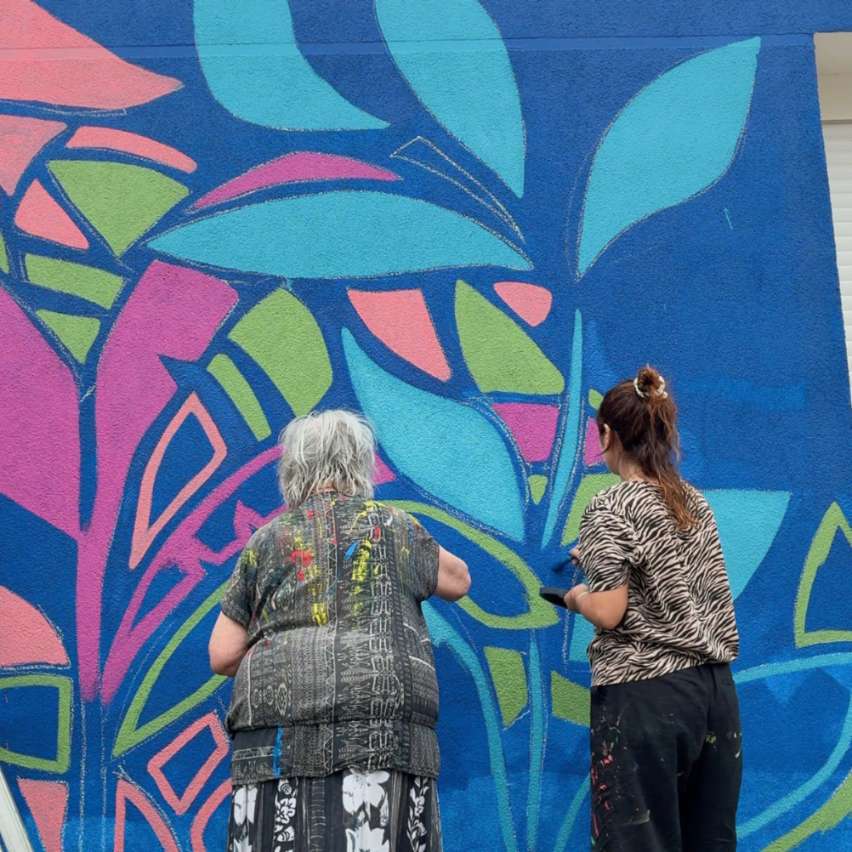 Deux femmes peignant une fresque murale végétale et colorée sur le mur extérieur d'une maison